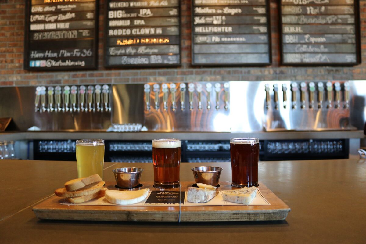 A flight of three craft beers with bread and cheese on a bar counter, with taps and a beer list behind