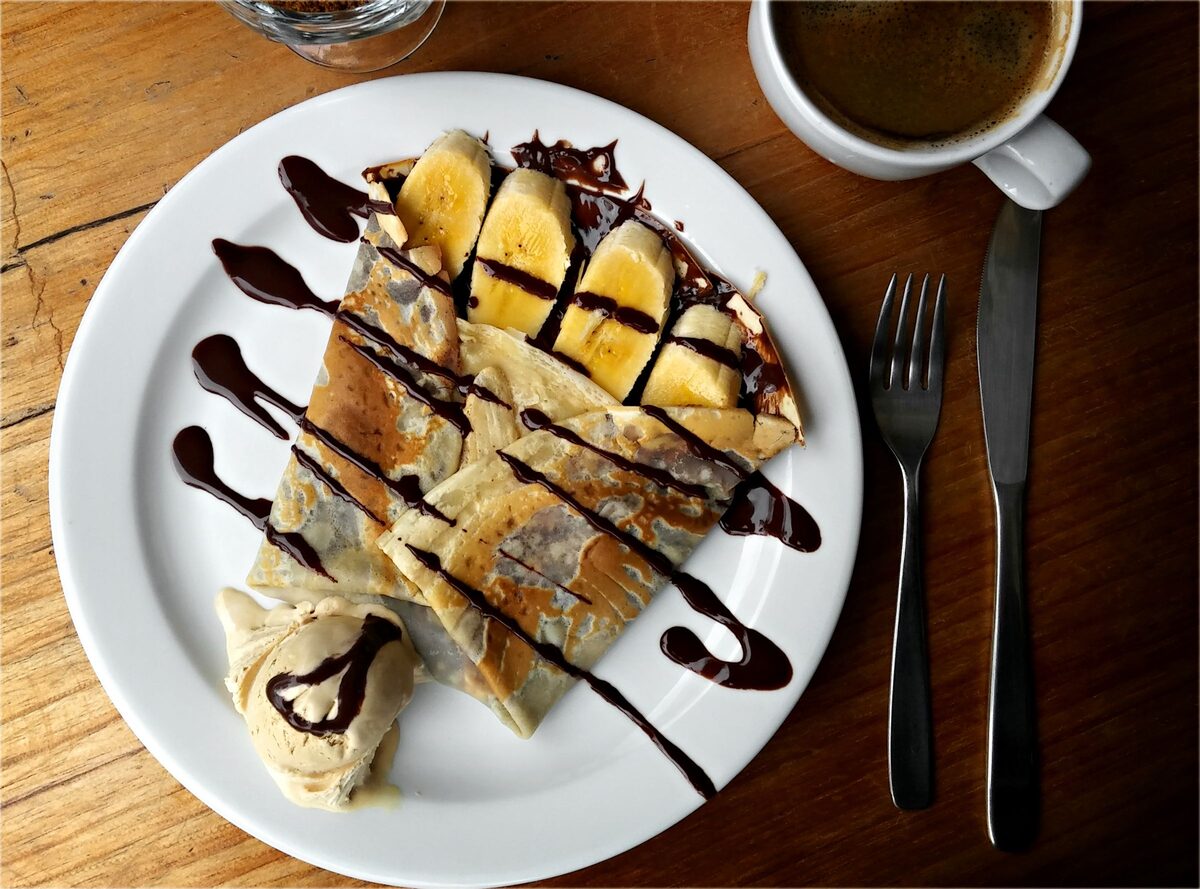 A folded crepe with banana slices and chocolate sauce, a scoop of ice cream and a cup of coffee on a wooden table