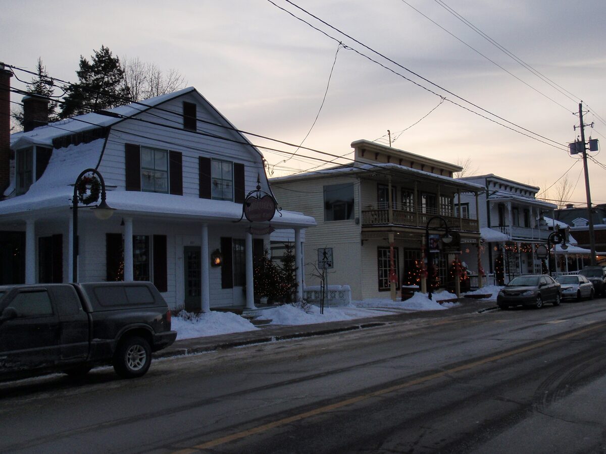 Snow-edged old wooden houses with porches and Christmas lights along a main street in Saint-Jovite at dusk