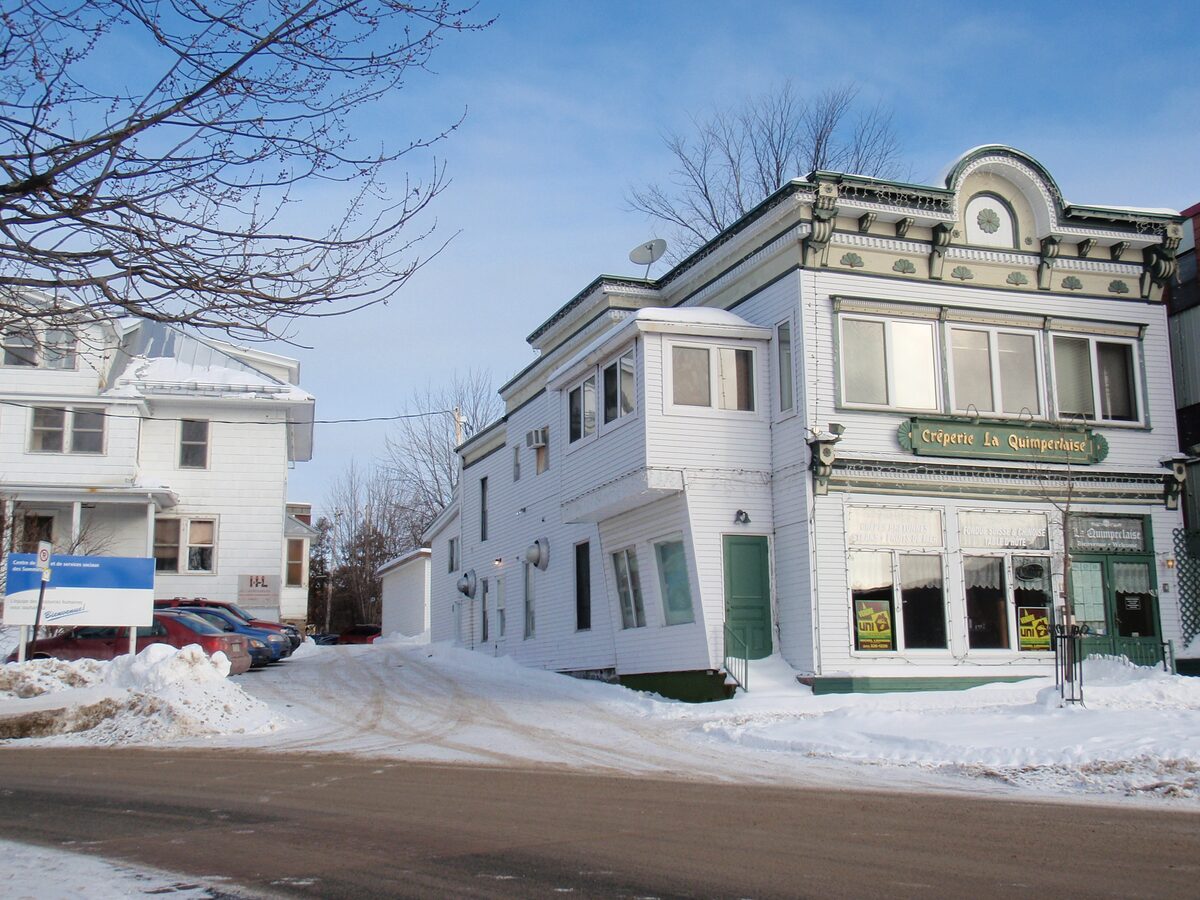 A white wooden corner building beside a snowy street in Sainte-Agathe-des-Monts on a sunny winter day
