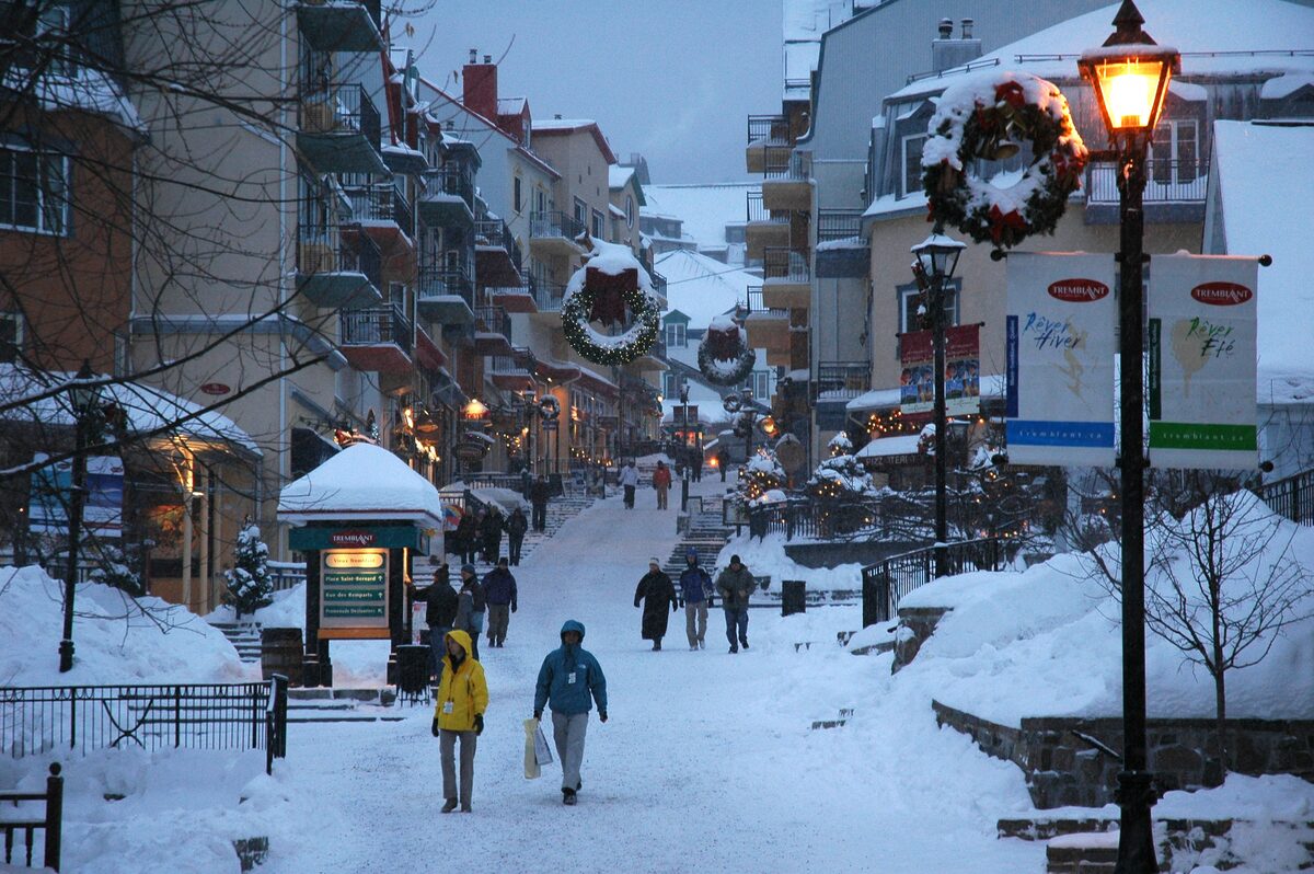 A snowy pedestrian lane in the Mont-Tremblant village at dusk, with wreaths on the lampposts and people walking