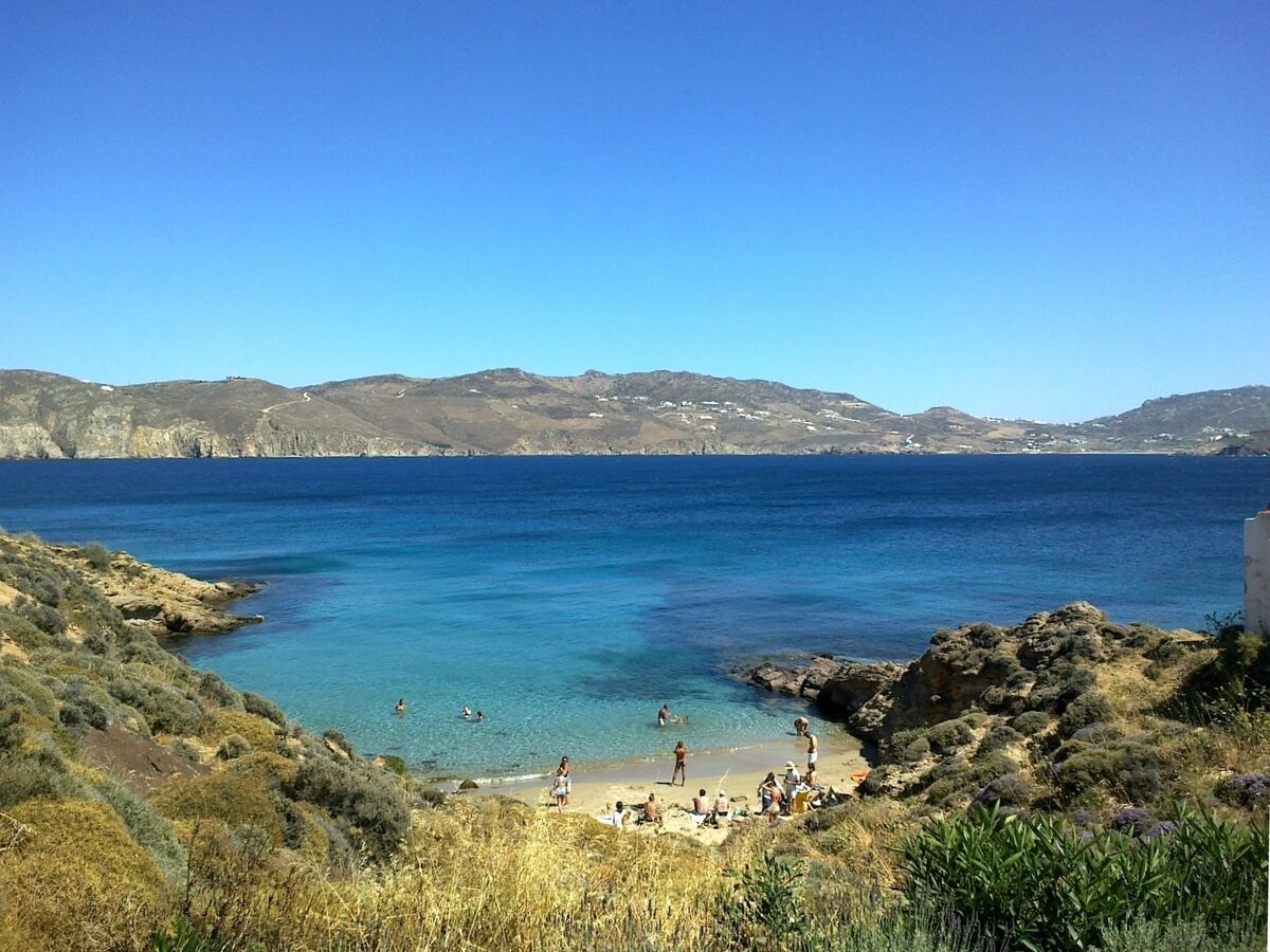 A small sandy cove with a few swimmers and clear blue water, backed by dry scrub and a long ridge across the bay