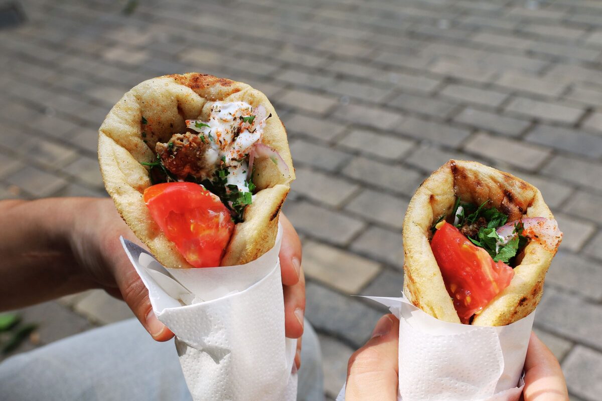 Two gyros wrapped in paper, held in hand over a paved street, with tomato and herbs in the pita
