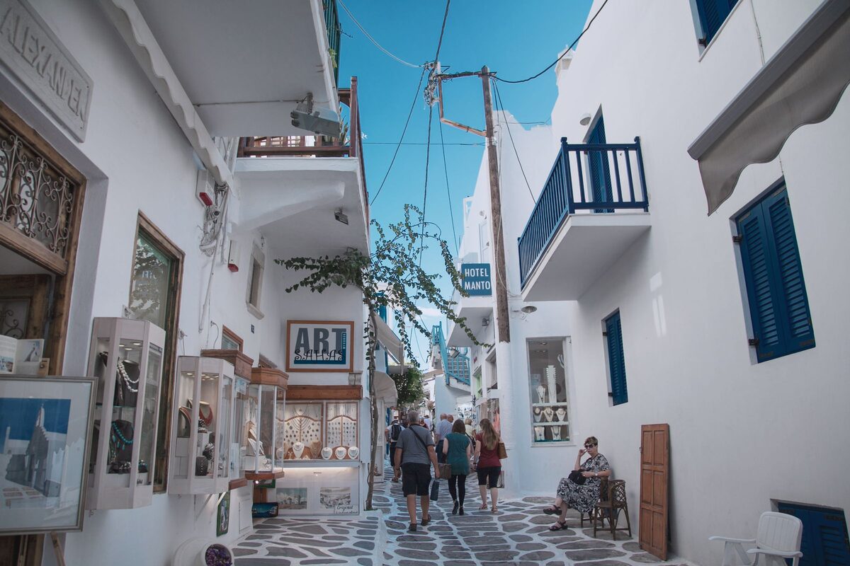 A whitewashed lane in Mykonos Town lined with shops and small galleries, with visitors walking down the paving