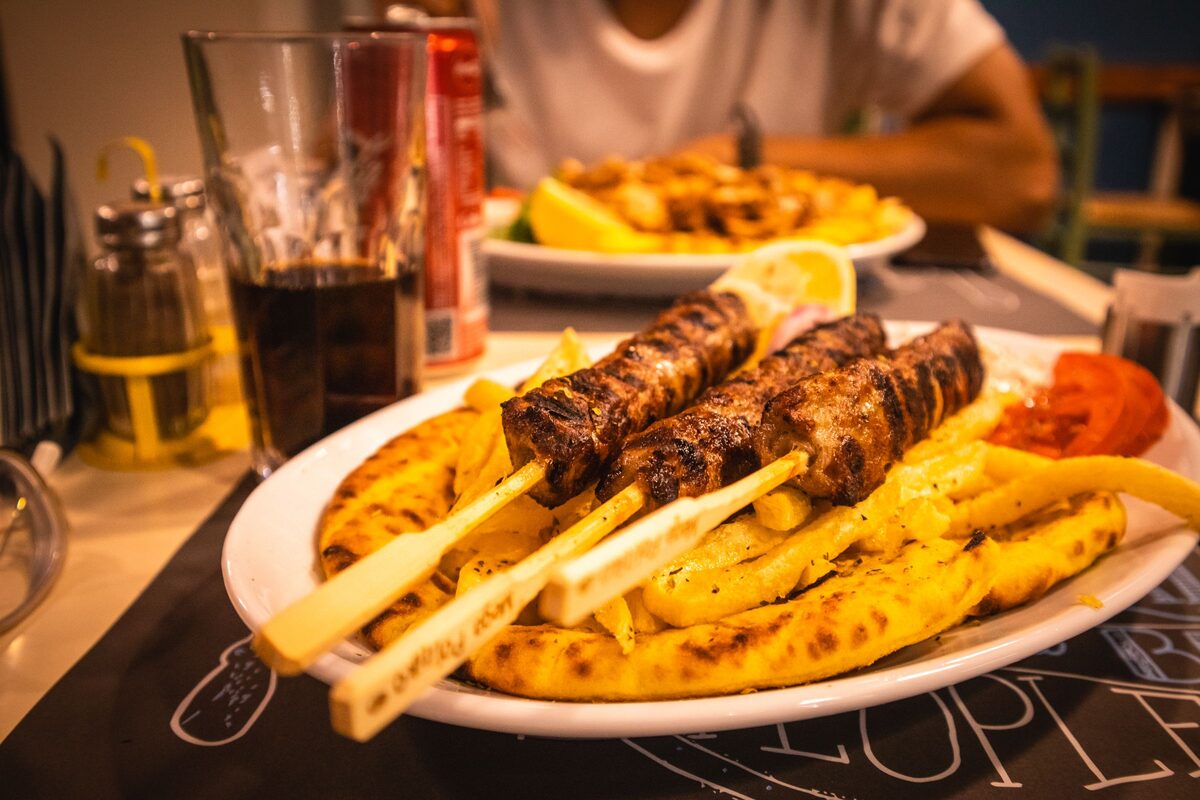 Grilled meat skewers on a plate of pita and fries, with a lemon wedge, on a taverna table