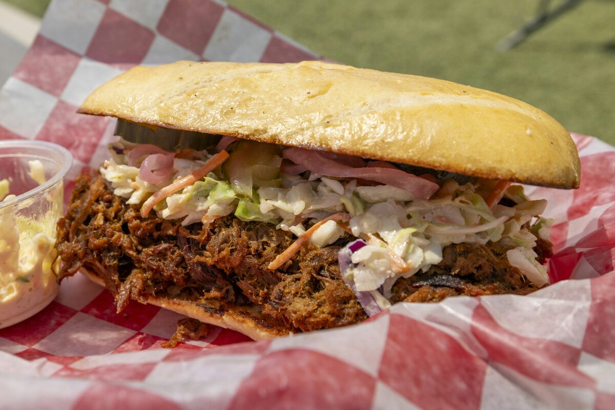A pulled pork sandwich with coleslaw and pickles on a red checkered paper liner