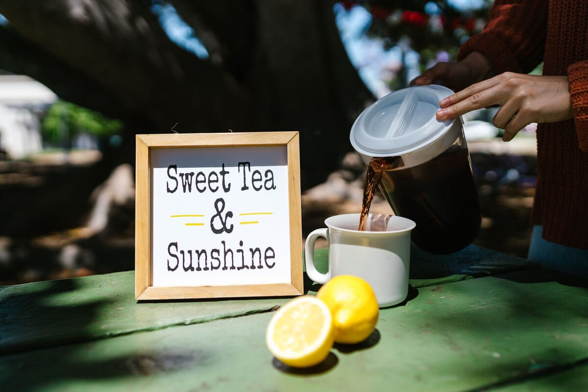 Dark tea poured from a pitcher into a mug beside a sweet tea sign and lemons