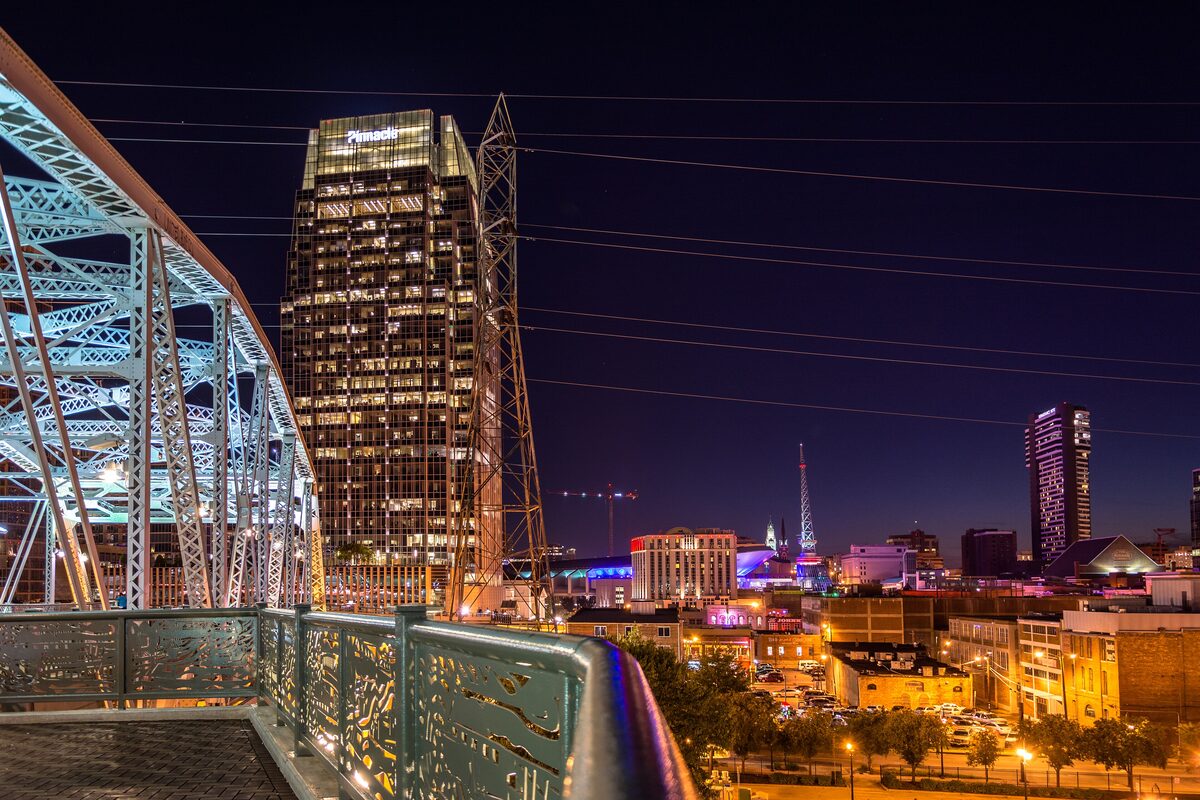 The lit steel trusses of the Seigenthaler Pedestrian Bridge at night above the downtown skyline