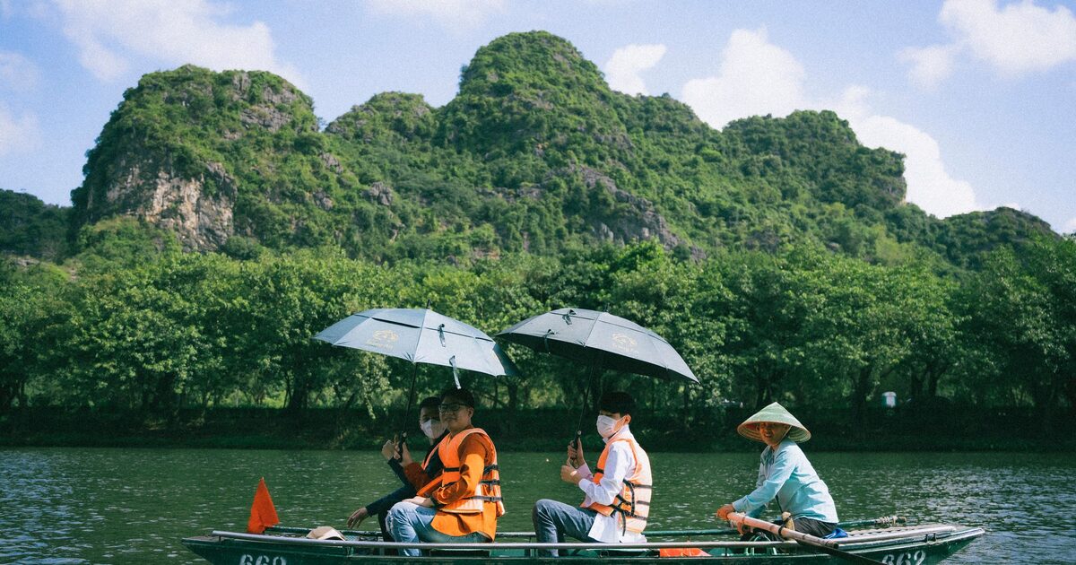 A Trang An rowing boat with passengers under umbrellas, below forested karst peaks