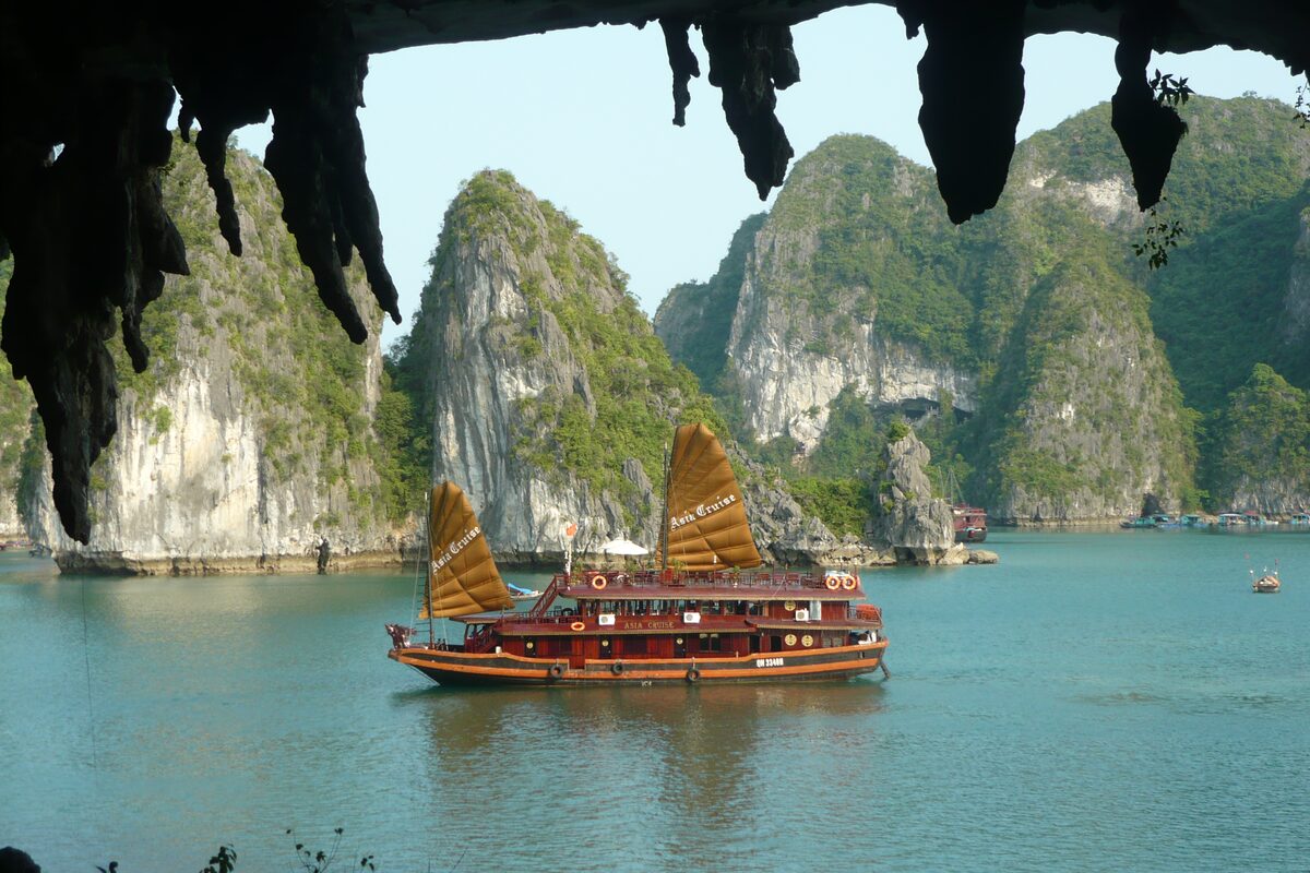 A traditional wooden junk sailing past limestone islands in Ha Long Bay, seen from under a cave overhang