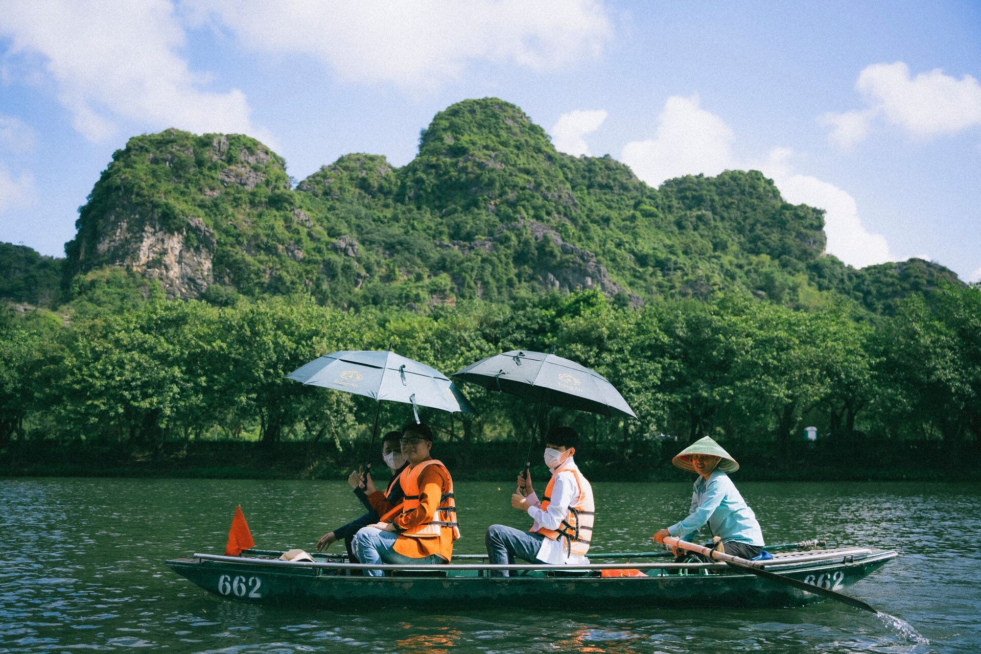 A Trang An rowing boat with passengers under umbrellas, below forested karst peaks
