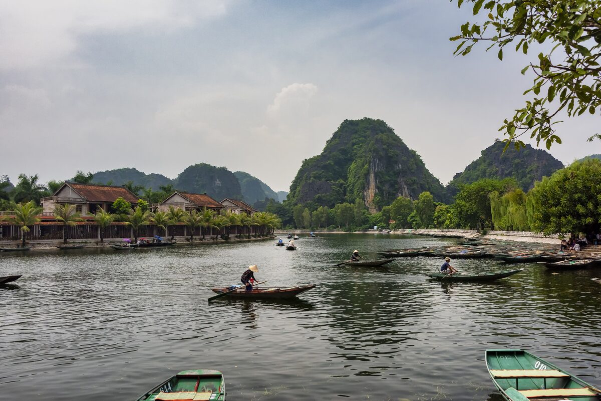 Wooden boats moored at the river wharf of Tam Coc, with village houses and karst peaks behind