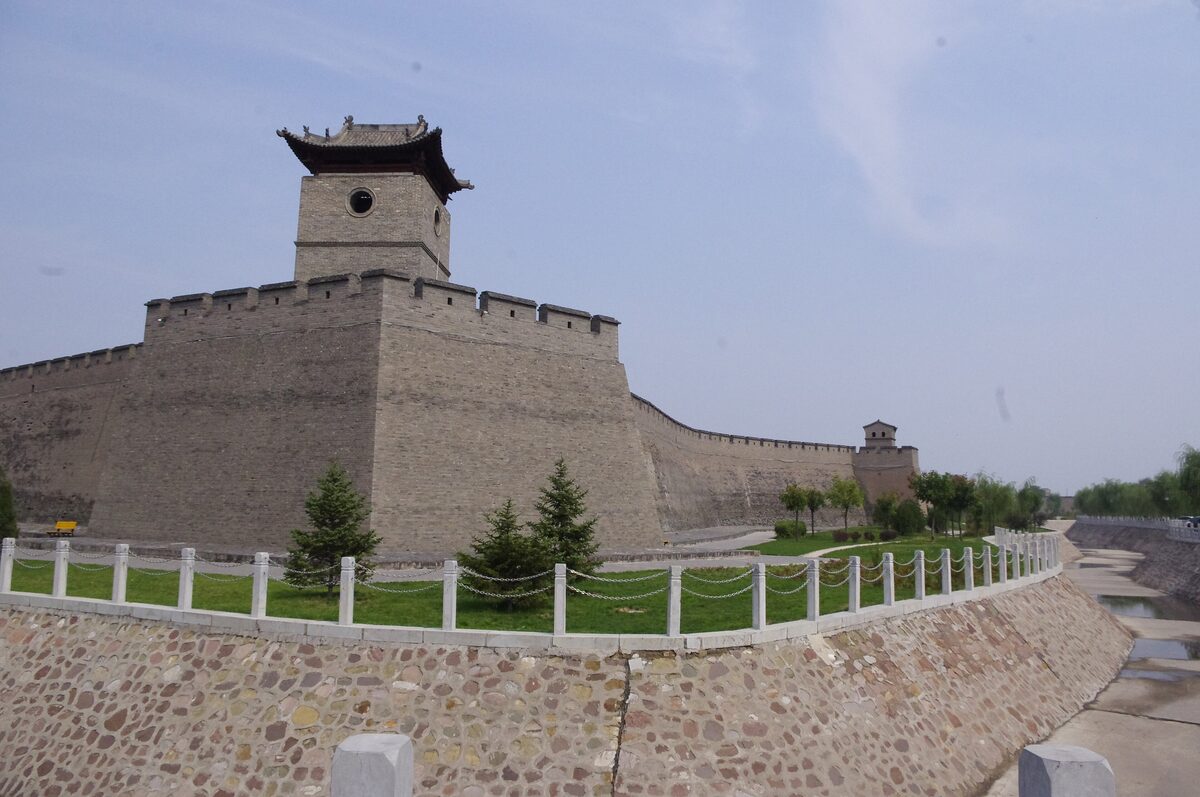 A corner watchtower on the grey brick Pingyao city wall, with a fenced lawn in front and a hazy sky behind