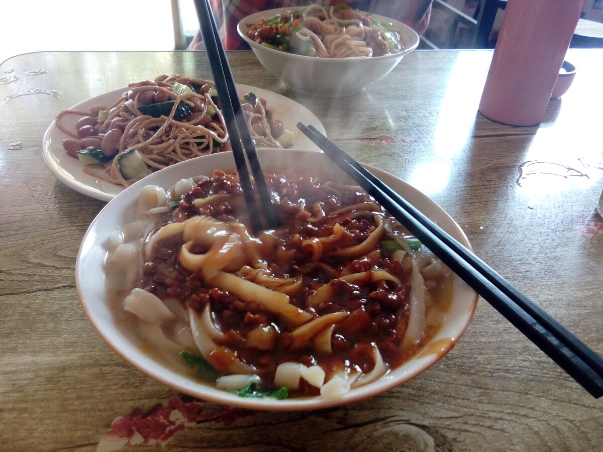 A bowl of knife-cut noodles in dark meat sauce with chopsticks, next to a second plate of noodles