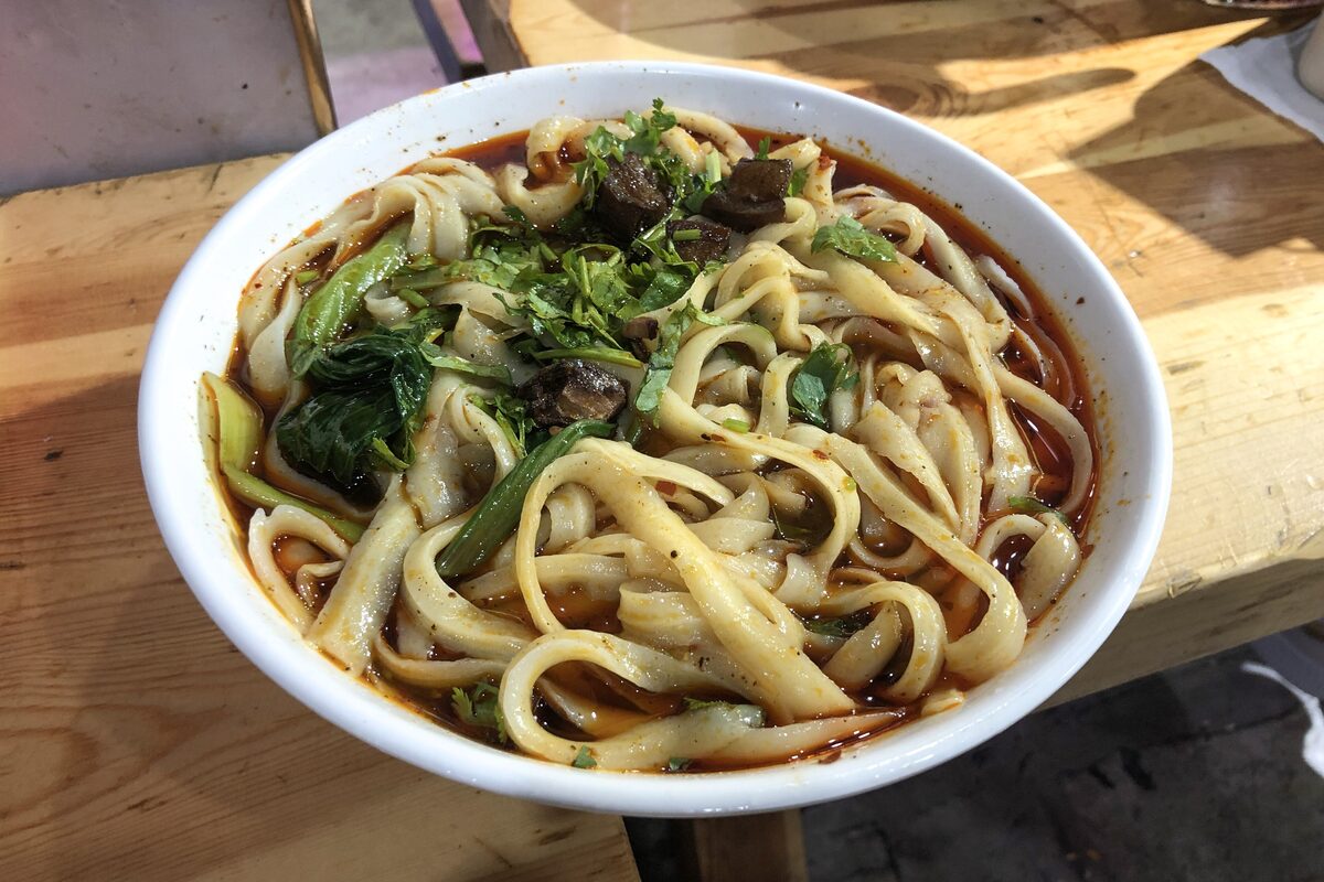 A white bowl of knife-cut noodles in red chili oil with greens and beef on a wooden table
