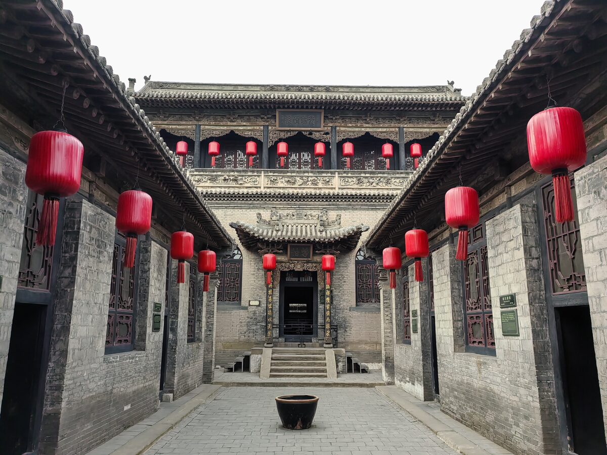 A narrow paved courtyard at the Qiao Family Courtyard lined with grey brick walls and red lanterns, with a gate hall at the end