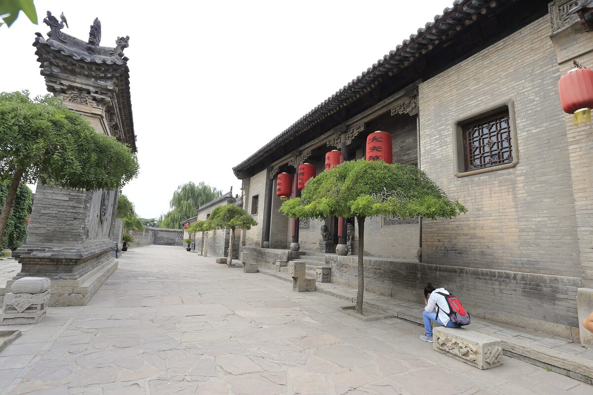 A wide stone-paved lane between grey brick courtyard walls at the Wang Family Compound, with red lanterns and a stone structure at the left