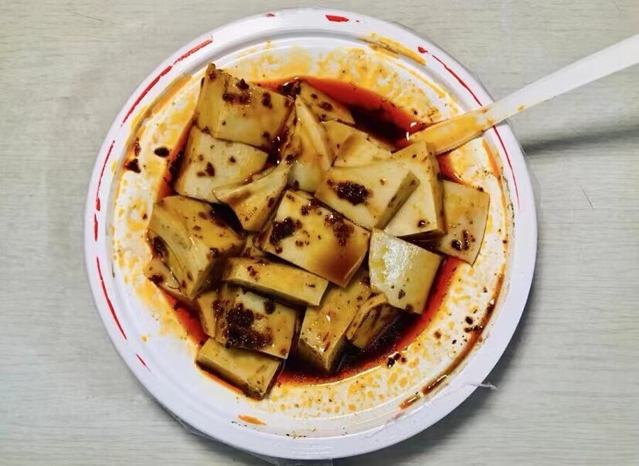 A paper bowl of wantuo, soft cut squares covered in red chili oil, with a spoon