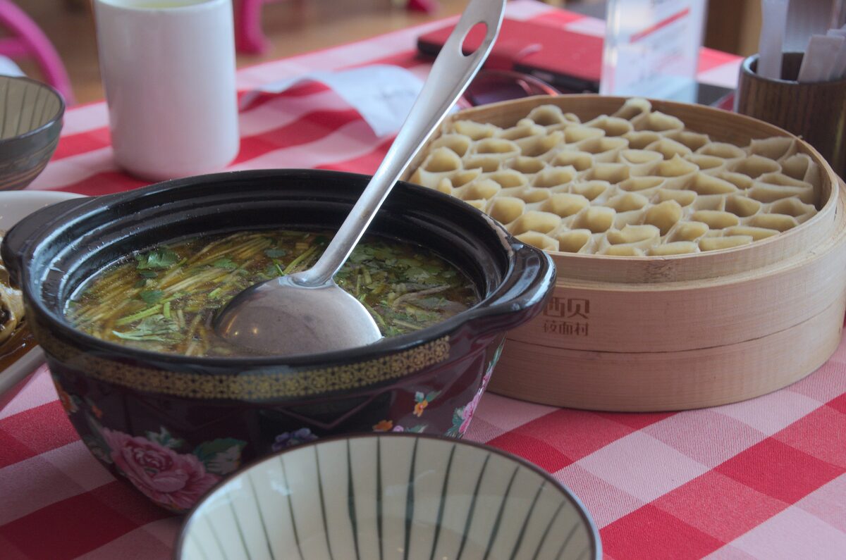 A steamer basket of oat-flour noodle pieces beside a clay pot of broth on a checked tablecloth