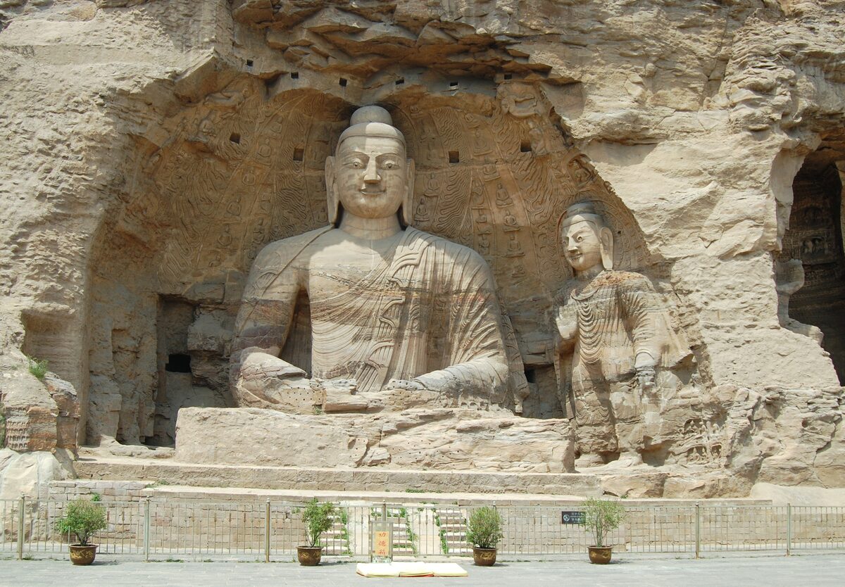 A seated Buddha carved into the sandstone cliff at the Yungang Grottoes, with a standing figure beside him