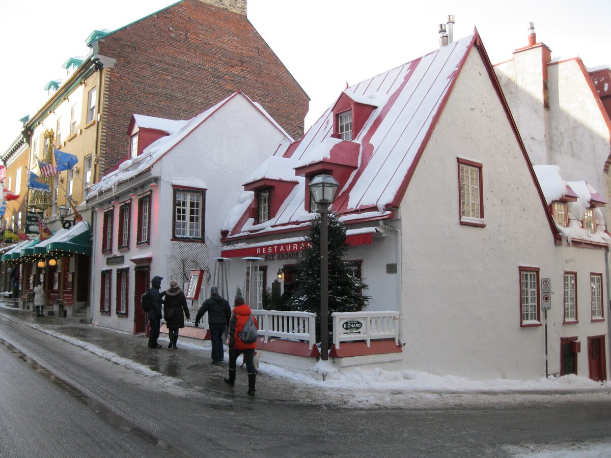The white 1675 house of Aux Anciens Canadiens with its red steep roof and snow on the street corner