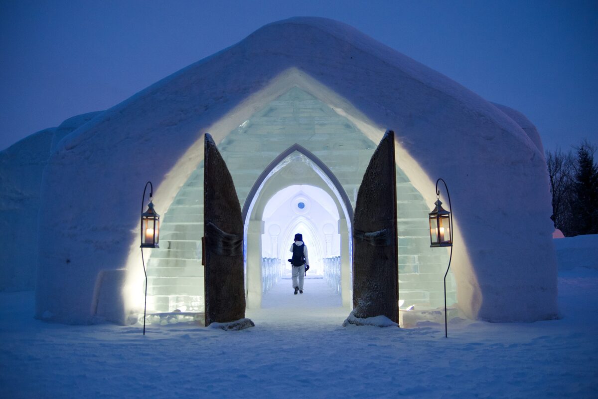 The snow-and-ice chapel entrance of an Hôtel de Glace lit by lanterns at dusk, with a visitor walking in