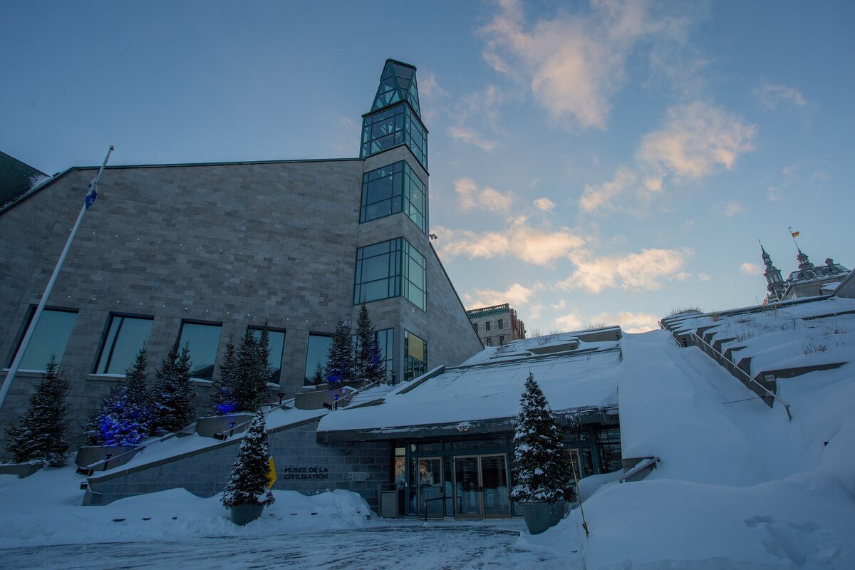 The stone and glass Musée de la civilisation at dusk in winter, with snowbanks and lit fir trees in front