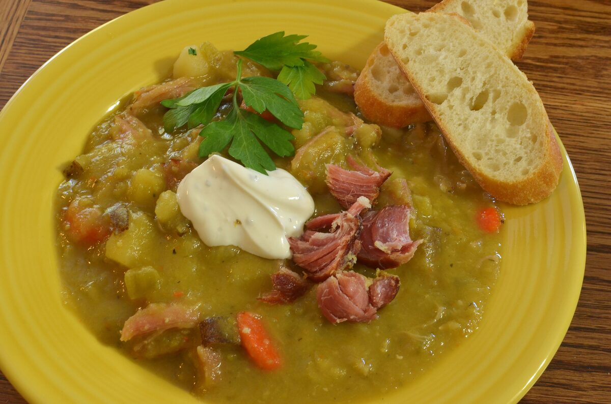A yellow bowl of thick split pea soup with pieces of ham, a spoon of cream and slices of bread