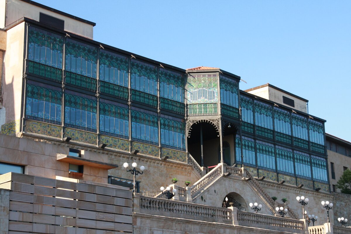 The southern facade of Casa Lis by day, a long stained-glass gallery above a stone terrace and a stair, under a blue sky