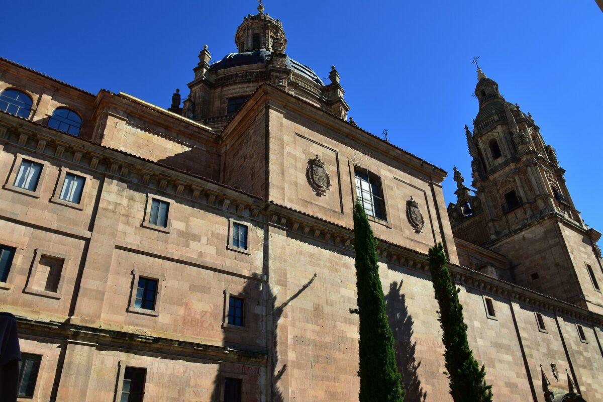 The sandstone side of La Clerecía with its dome and baroque bell tower, two cypresses in front, under a deep blue sky