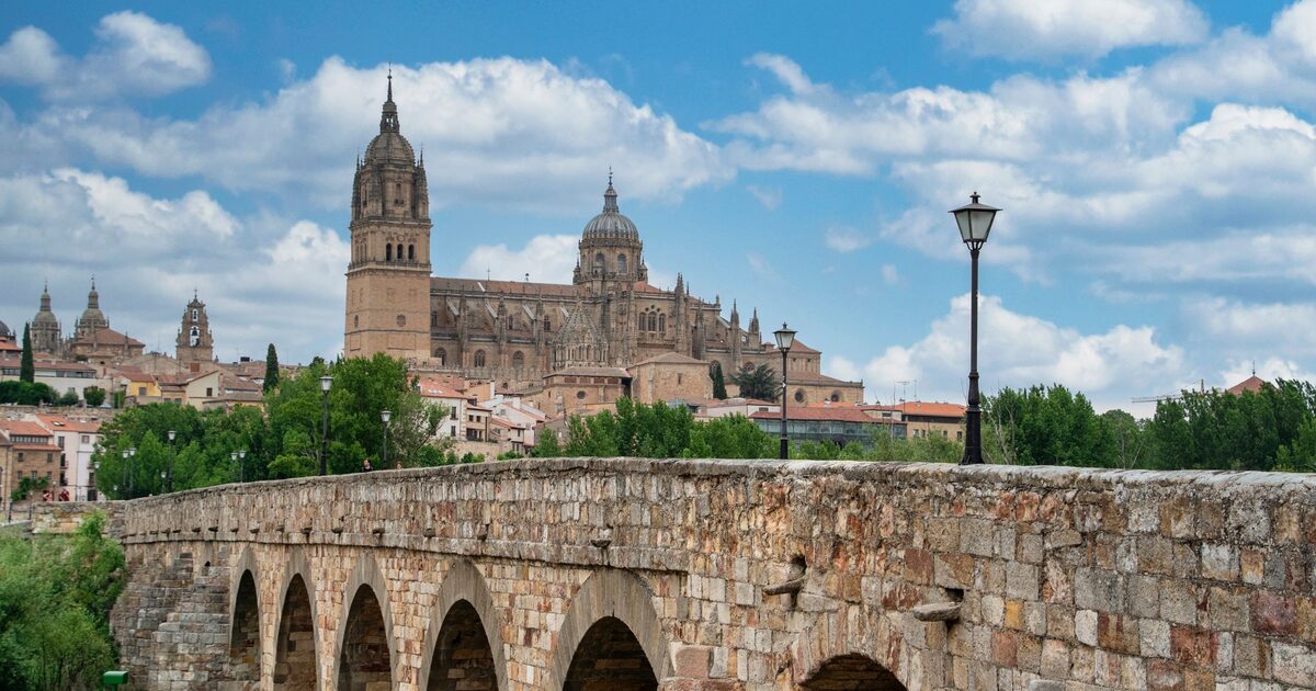 The New Cathedral and its tall tower above the old town, seen over the stone arches of the Roman bridge under a blue sky