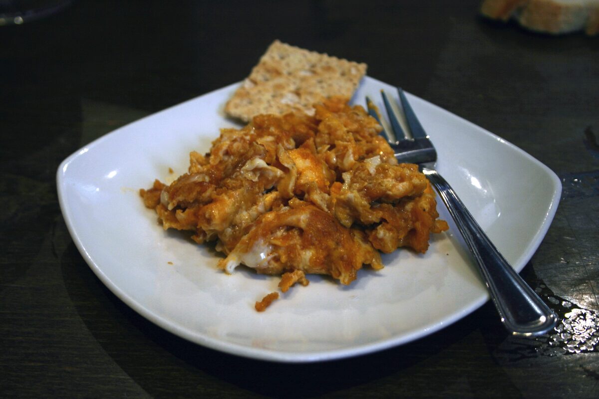 A white plate of paprika-orange scrambled eggs with farinato, a cracker and a fork on the side