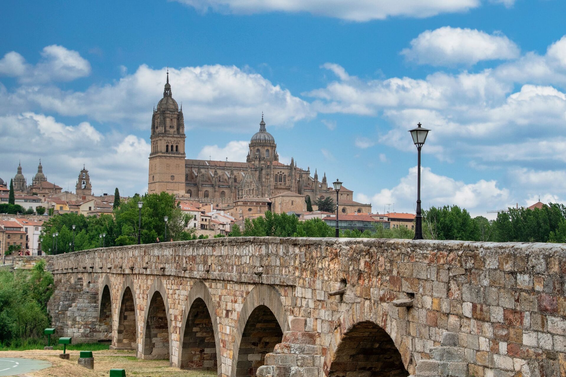 The New Cathedral and its tall tower above the old town, seen over the stone arches of the Roman bridge under a blue sky