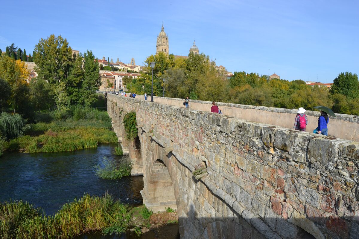 The side of the Roman bridge over the Tormes, with walkers on top, autumn trees and the cathedral tower beyond