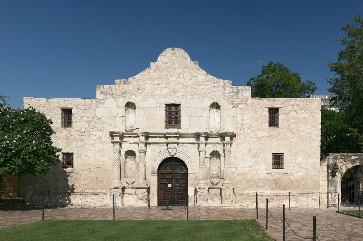 The weathered limestone facade of the Alamo church with its carved doorway and curved gable under a blue sky
