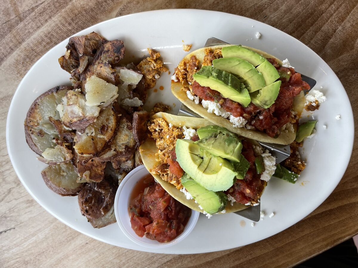 Two soft breakfast tacos with egg, avocado and salsa beside a pile of potatoes on a white plate