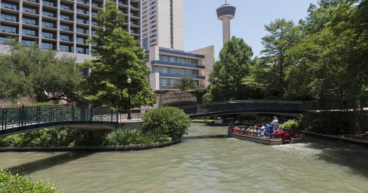 A red river barge with visitors passes under a footbridge on the San Antonio River Walk, with the Tower of the Americas rising behind the trees