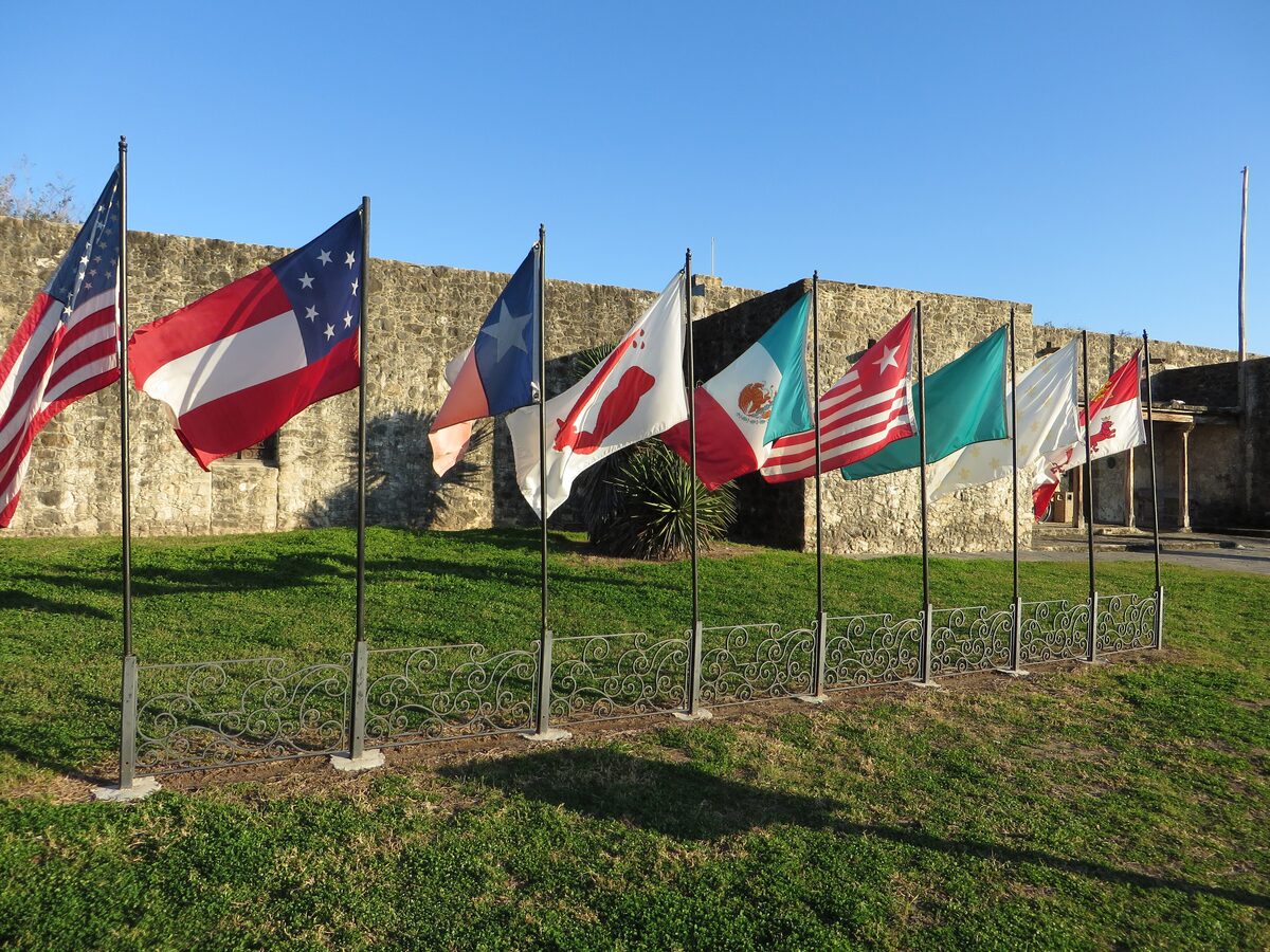 A row of flags on the lawn in front of the stone walls of Presidio La Bahía in Goliad