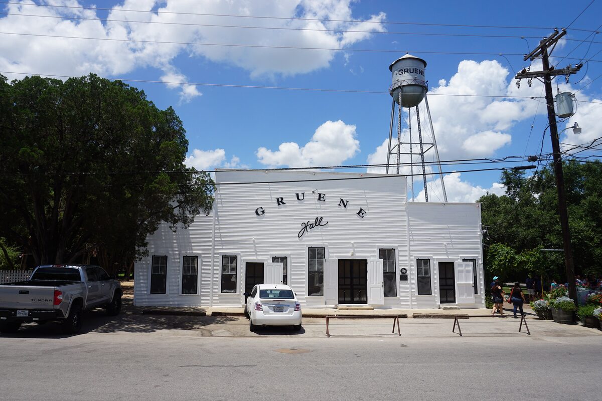 The white wooden Gruene Hall with its painted sign, a water tower behind it and flower pots by the street