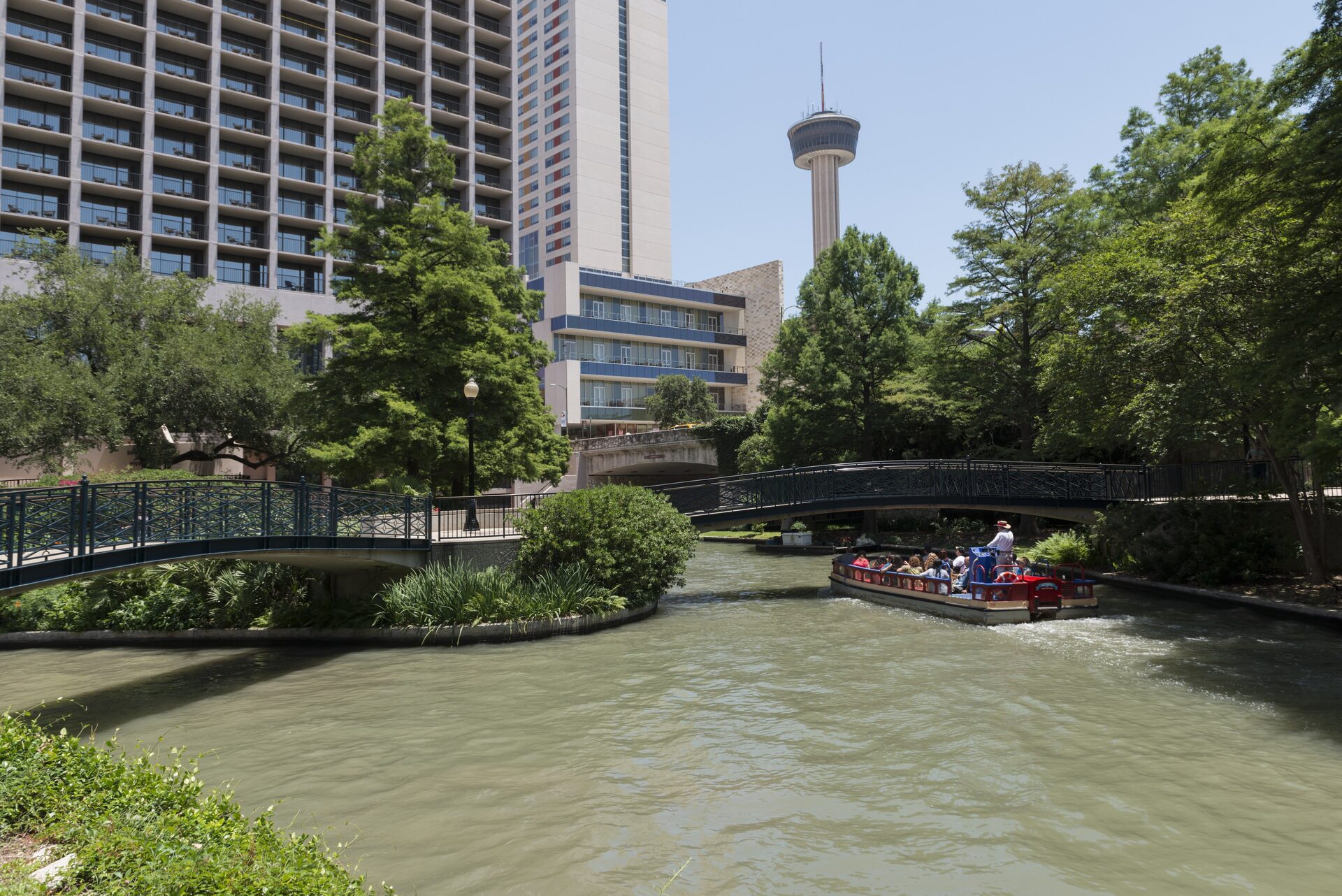 A red river barge with visitors passes under a footbridge on the San Antonio River Walk, with the Tower of the Americas rising behind the trees