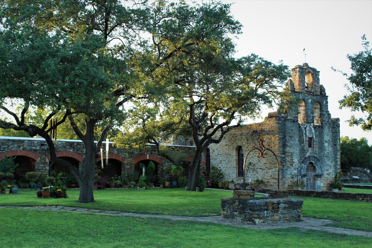 The small stone chapel and bell gable of Mission Espada behind a courtyard well and live oaks