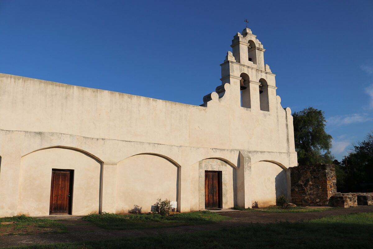 The whitewashed bell-gable chapel and arcade of Mission San Juan in warm light