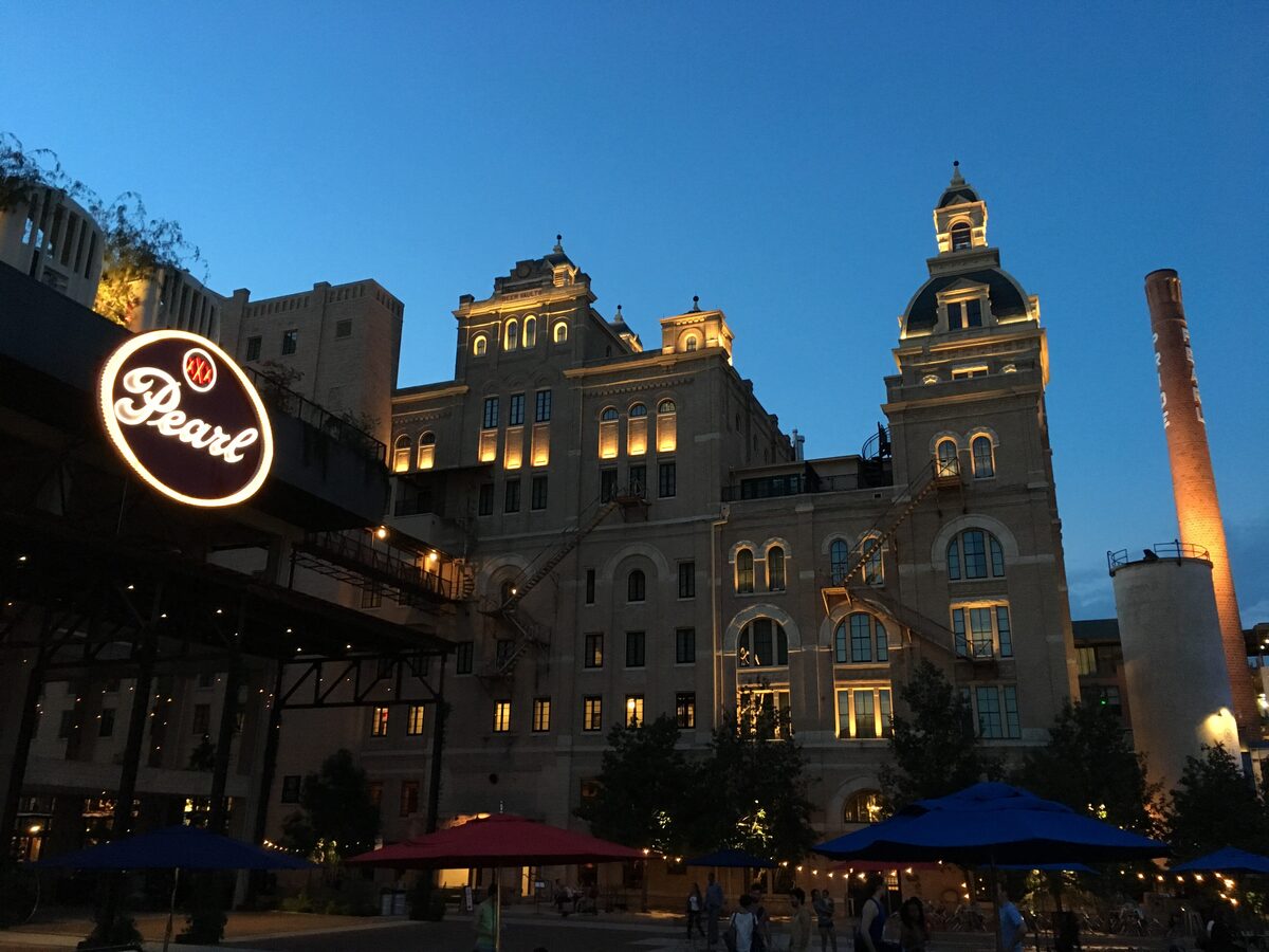 The lit-up Pearl brewhouse tower and glowing Pearl sign at dusk, with umbrella tables below