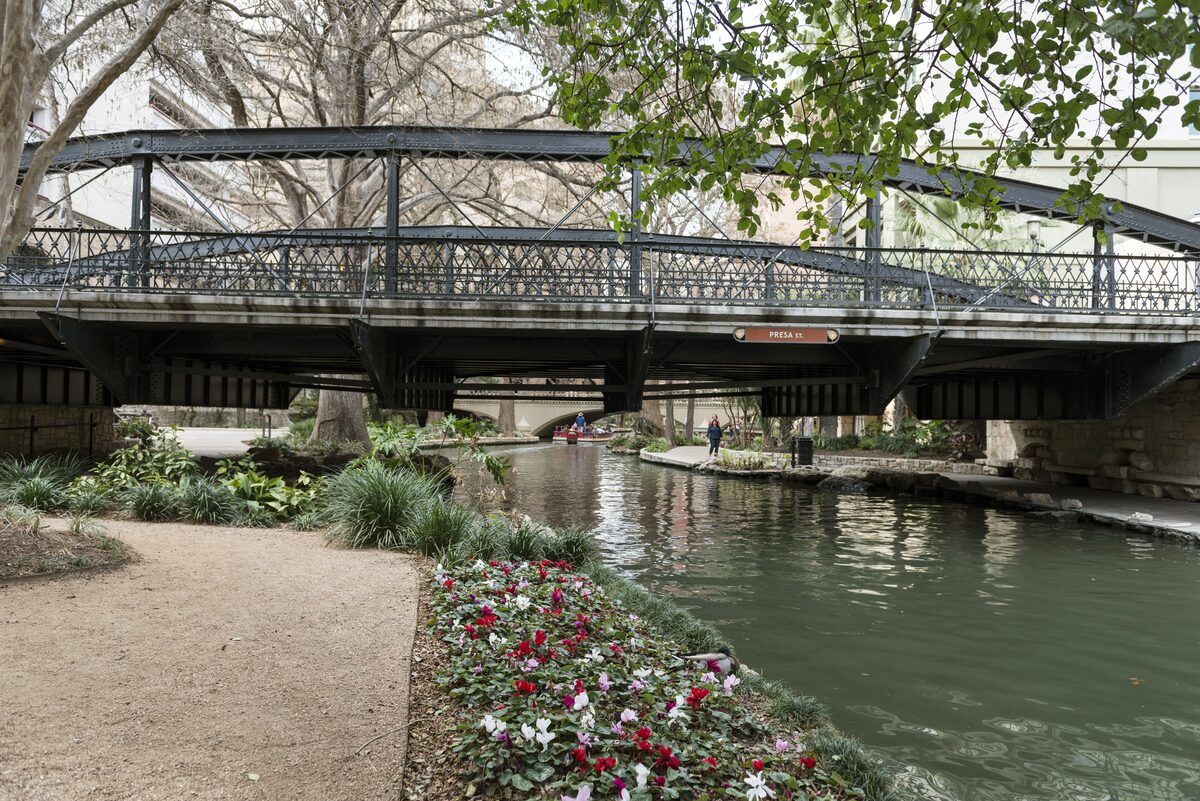 An iron-trussed bridge over the green San Antonio River with a flower bed along the walkway