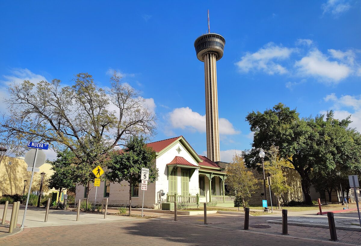 The Tower of the Americas rising above a small Victorian house and trees at Hemisfair under a blue sky