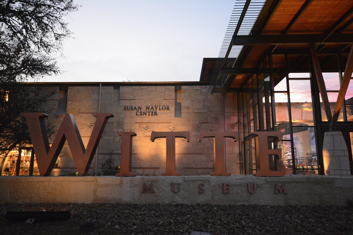 Large copper letters spelling WITTE in front of the Witte Museum's stone and glass entrance at dusk