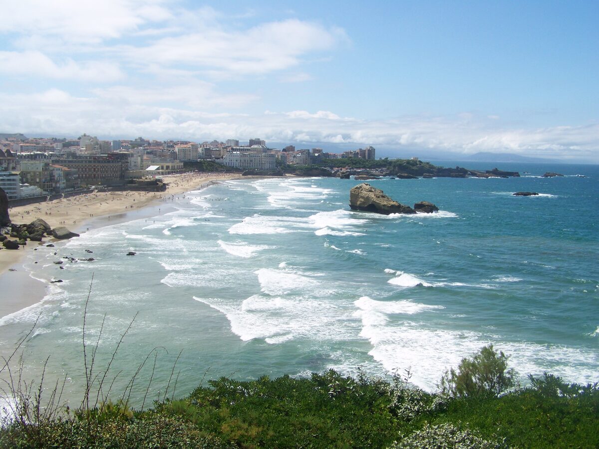 Biarritz beach from above, with white surf breaking across the sand and a rock stack standing in the sea