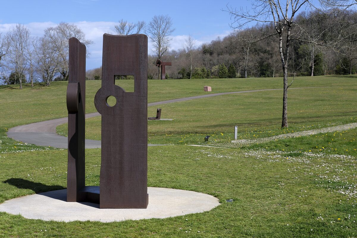 A tall steel sculpture by Chillida on a round pad in the grass of the park, with a path and bare trees behind