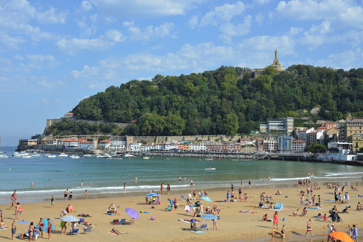 Playa de la Concha on a sunny day, the sand full of people, the sea in front and Monte Urgull with its statue at the far end