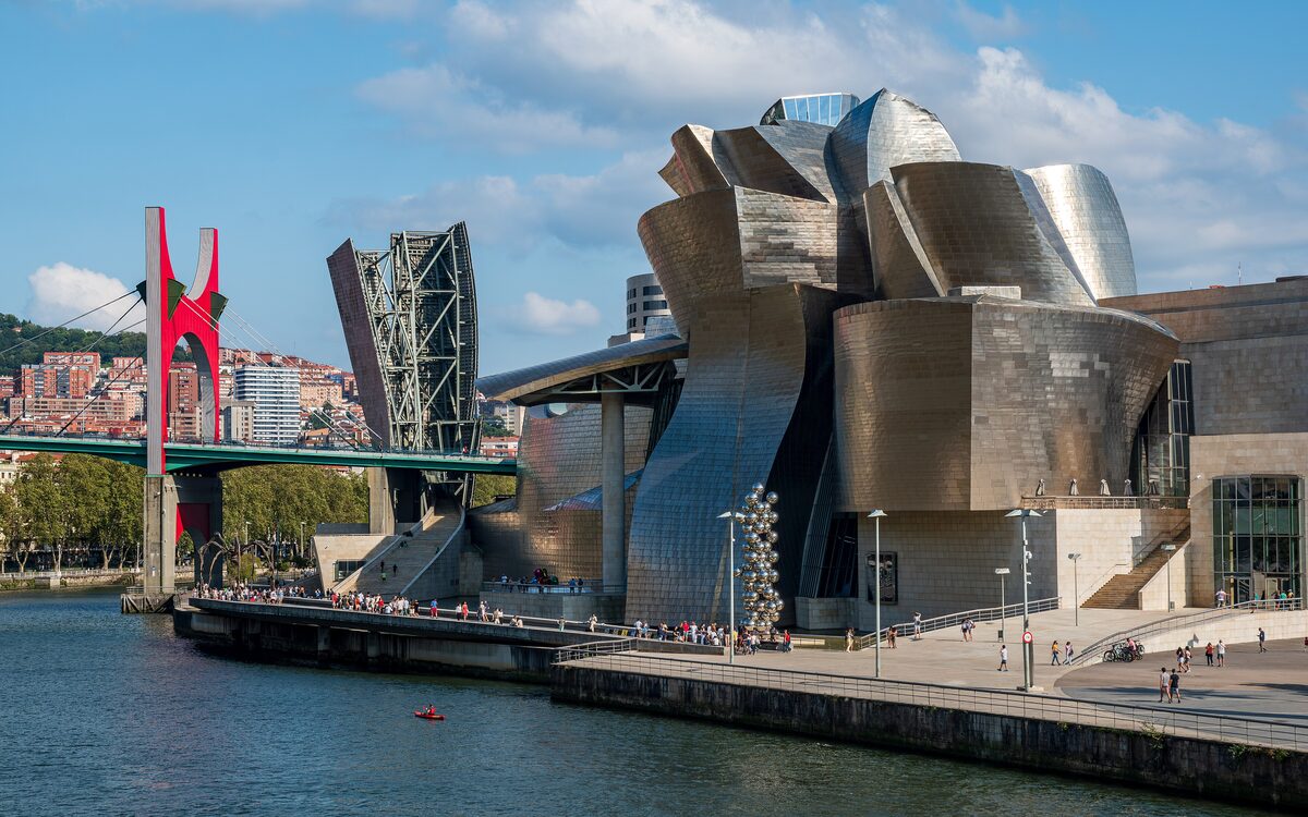 The Guggenheim Museum in Bilbao, its curved metal panels catching the light, with a red arch bridge over the river beside it